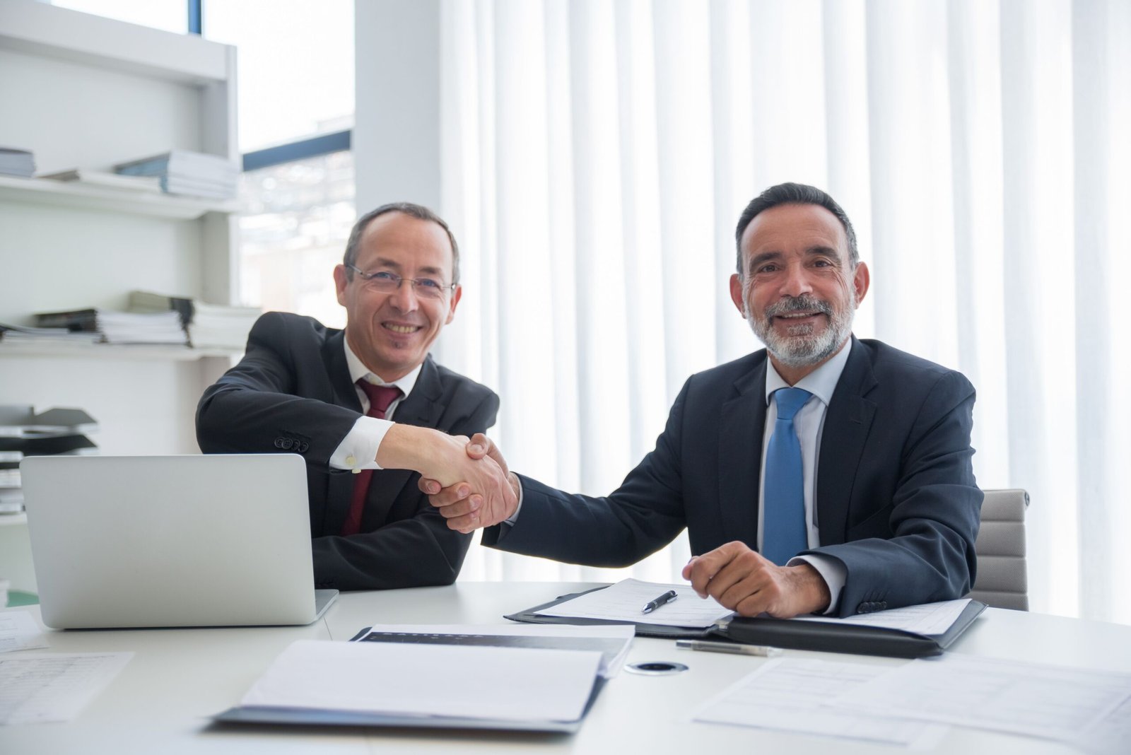 Two business professionals shaking hands with contract documents on the table, representing a supplier contract agreement